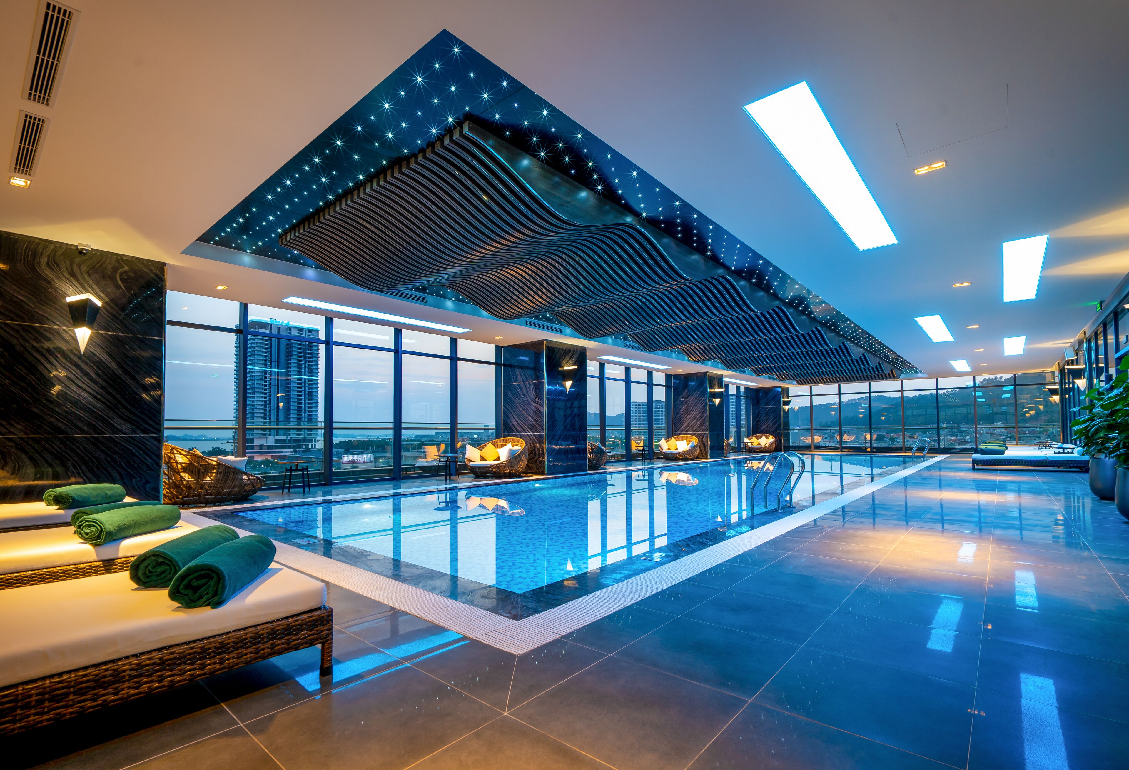 A modern indoor swimming pool with a high ceiling, large windows, and a lounge area with seating and towels.