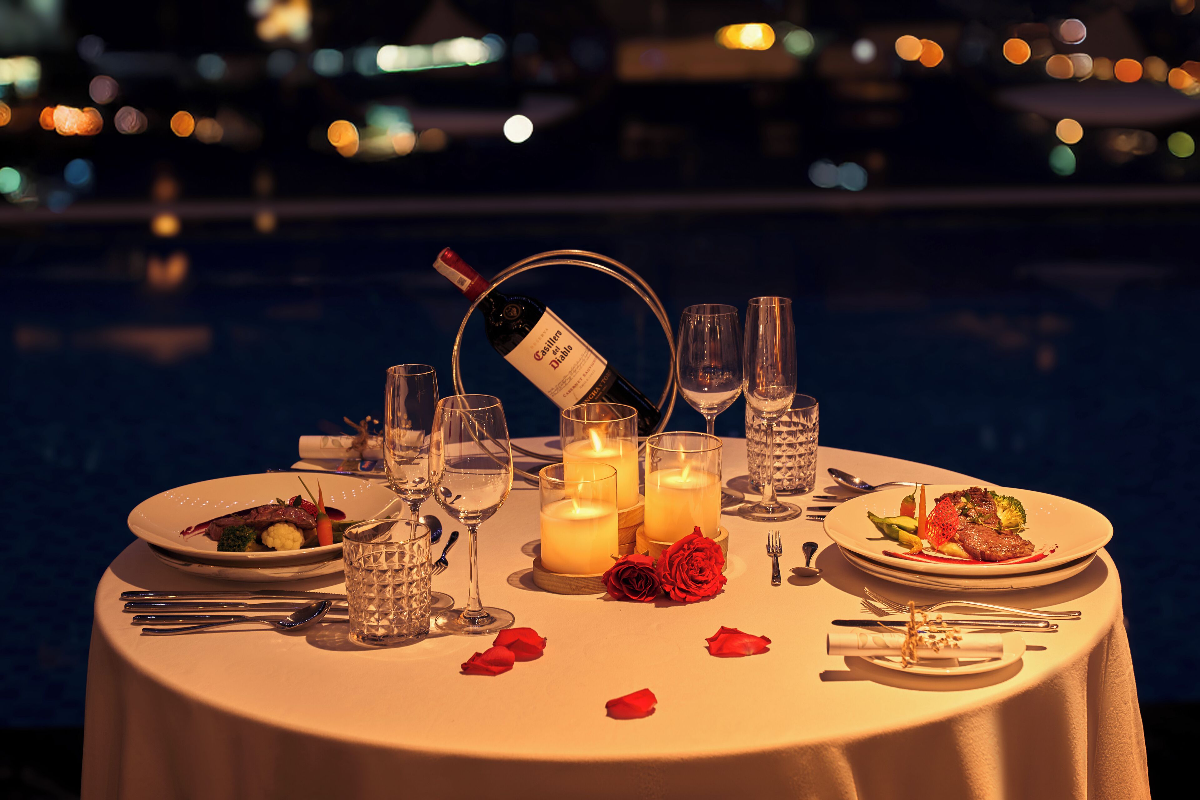 Una mesa dispuesta para una cena romántica, con velas, copas y pétalos de rosa.