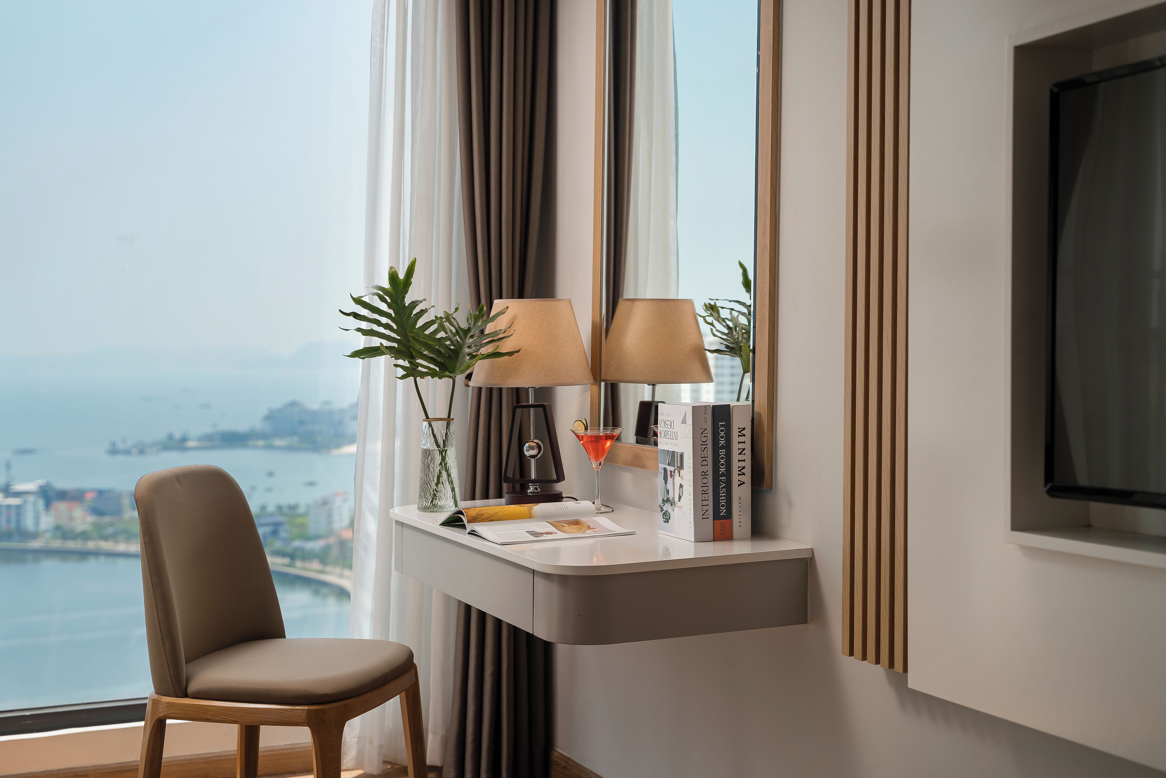 Habitación de hotel moderna con vista al mar, un escritorio con lámpara, una silla y una estantería.
