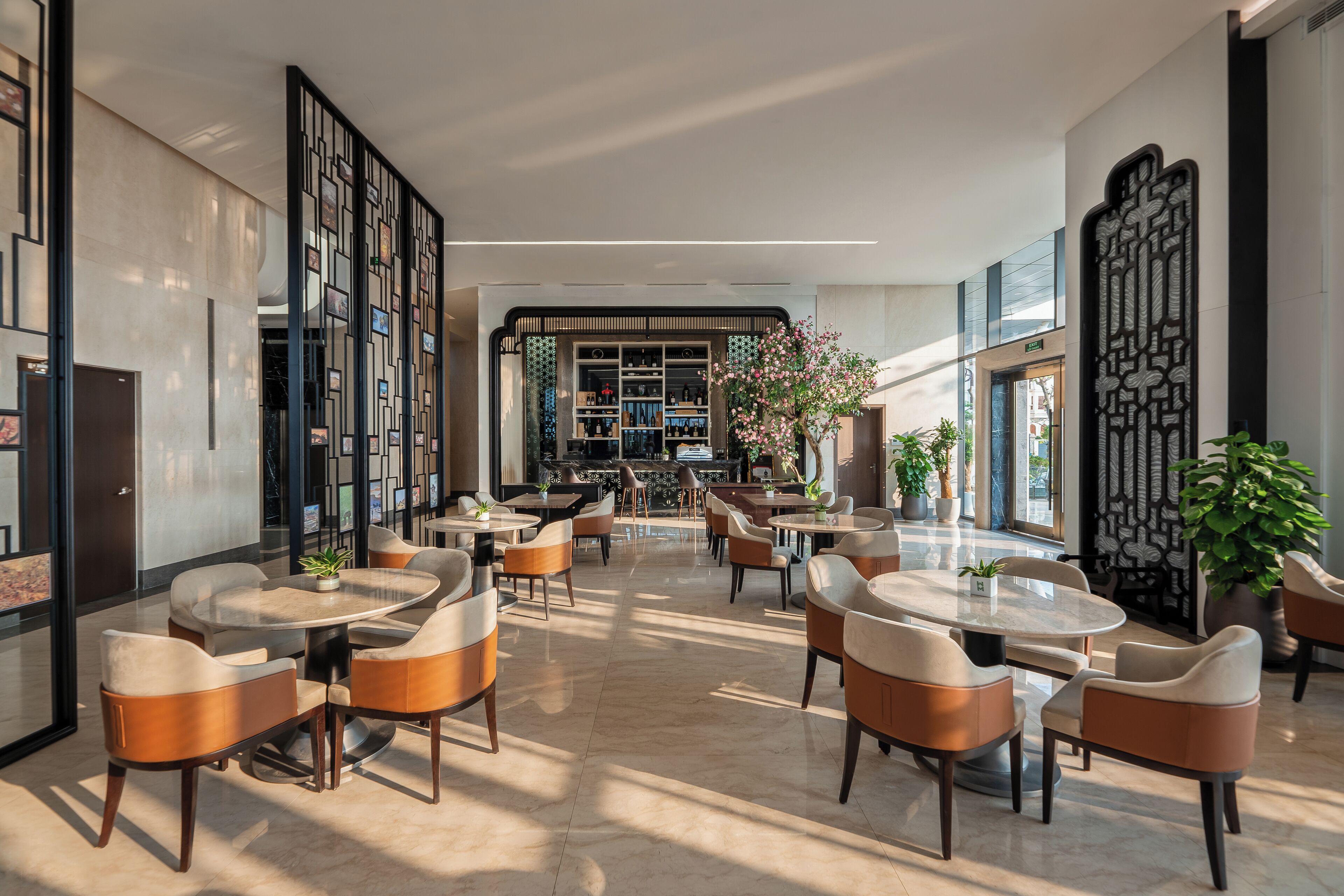 Un moderno hall de hotel con mesas y sillas redondas, una zona de bar central y paneles decorativos.