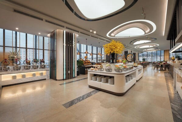 Un moderno vestíbulo de hotel con una isla central que presenta un arreglo floral, un área de descanso con mesas y un mostrador de buffet con diversos alimentos.