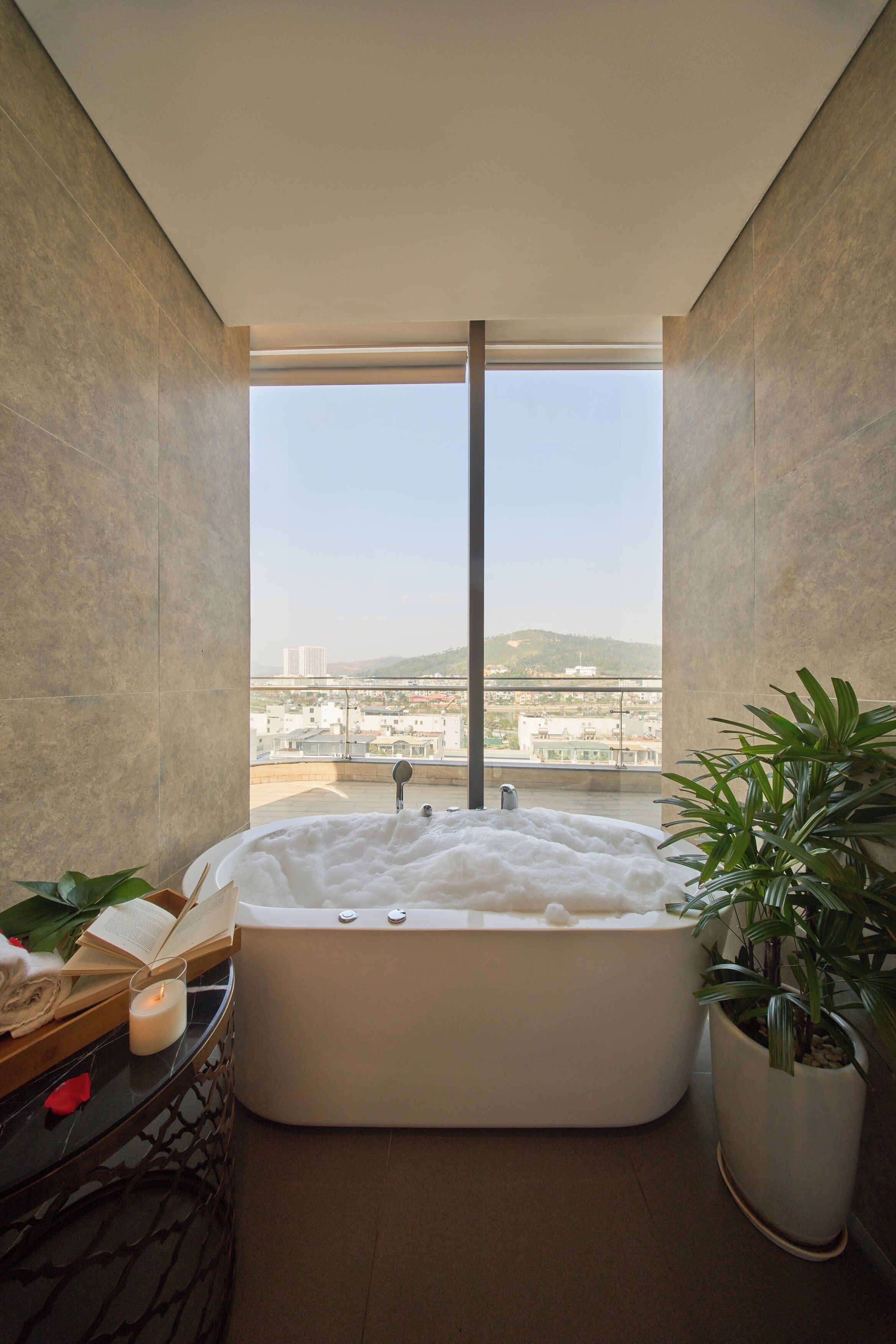 A modern bathroom with a large bathtub, a potted plant, and a view of a cityscape through a large window.