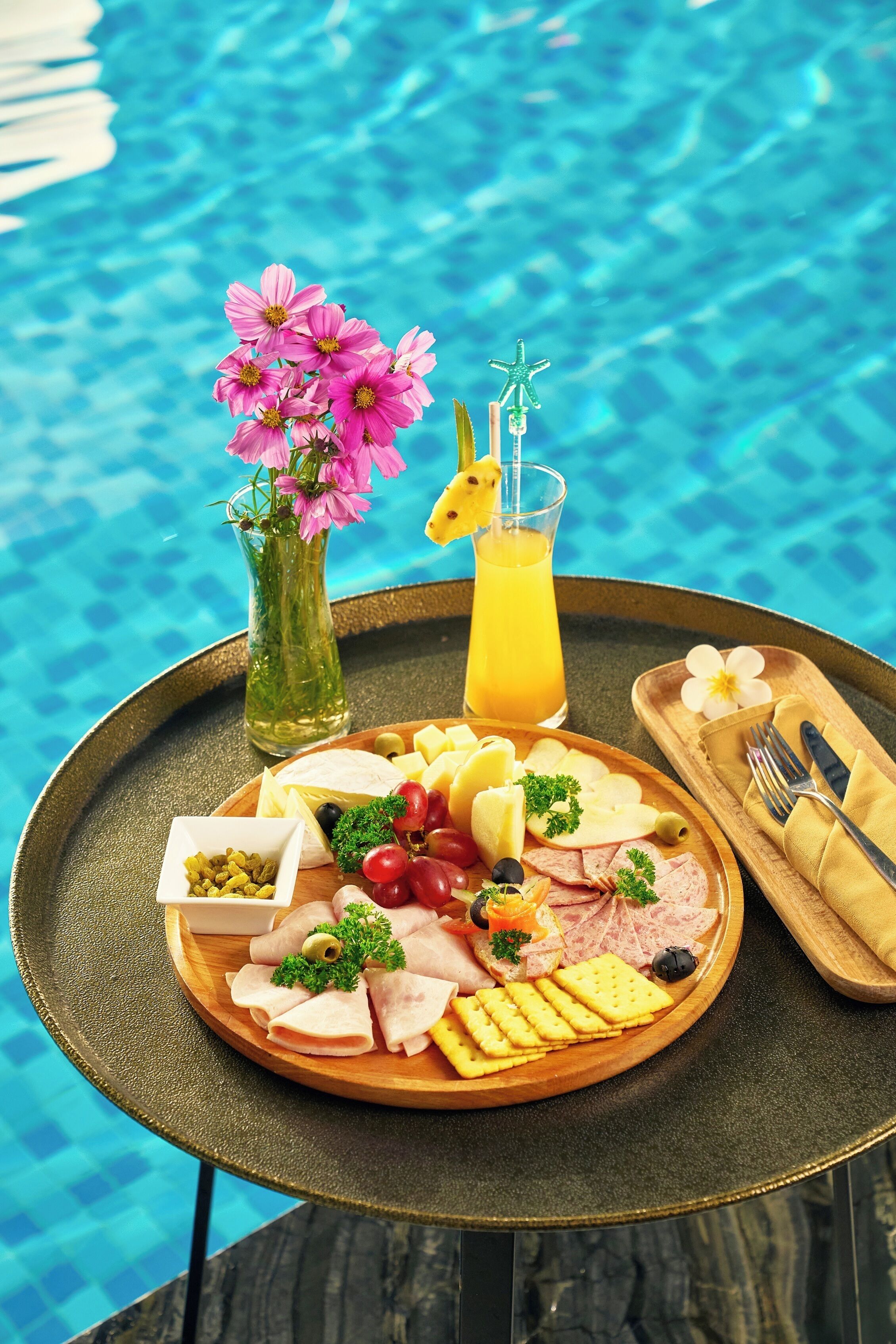 A tray with a variety of food items including cheese, fruits, and crackers, placed on a table by a poolside.