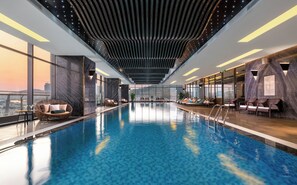 An indoor swimming pool with a view of the cityscape, modern lounge chairs, and a ceiling with a unique light design. - DeLaSea Ha Long Hotel (Ha Long)