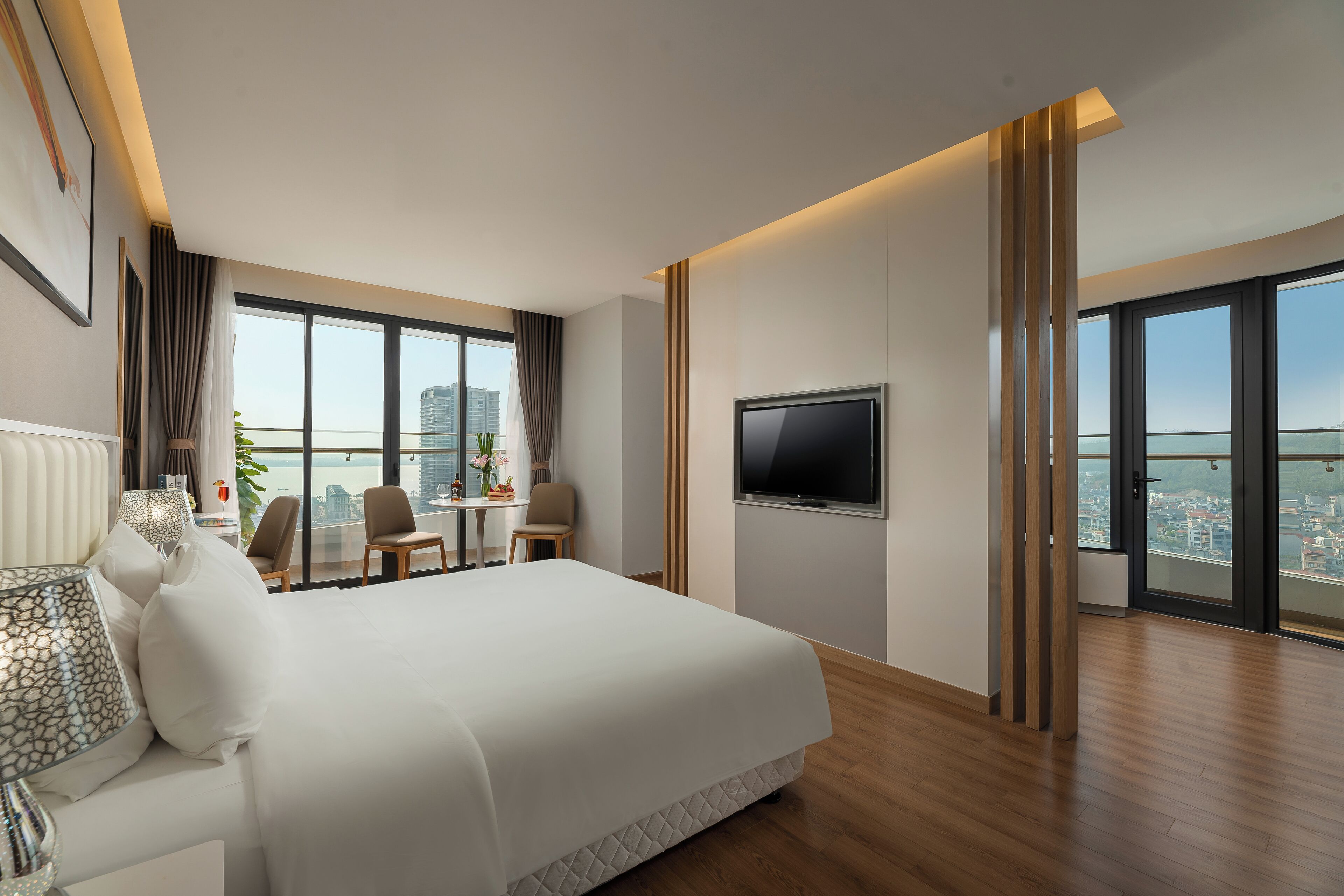 A modern hotel room with a large bed, a TV mounted on the wall, and a city view through large windows.