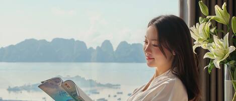 A person sitting by a window with a view of a body of water and distant landforms, reading a book.