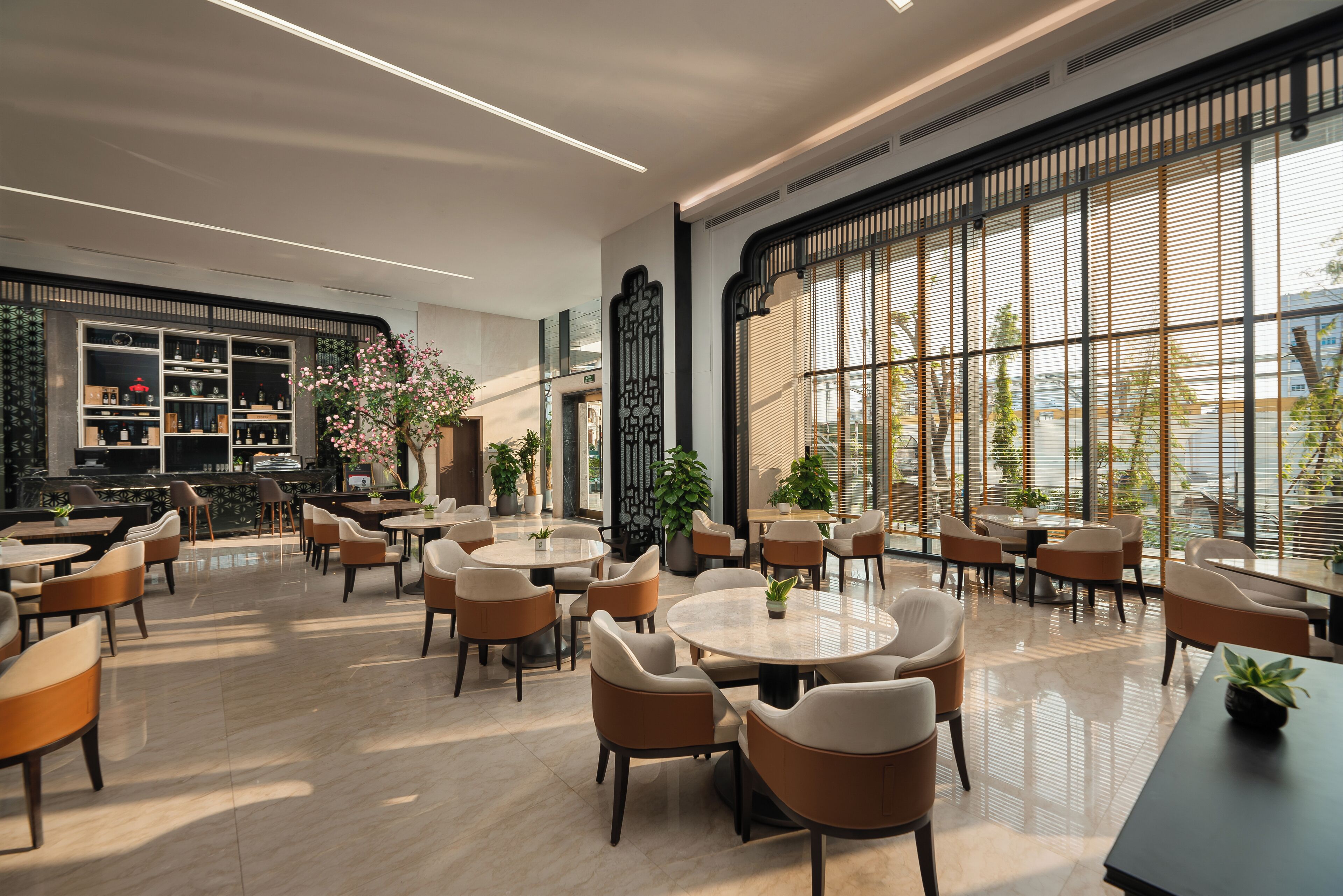 A modern indoor space with round tables and chairs, a bar area with shelves, and large windows offering views of the outside.