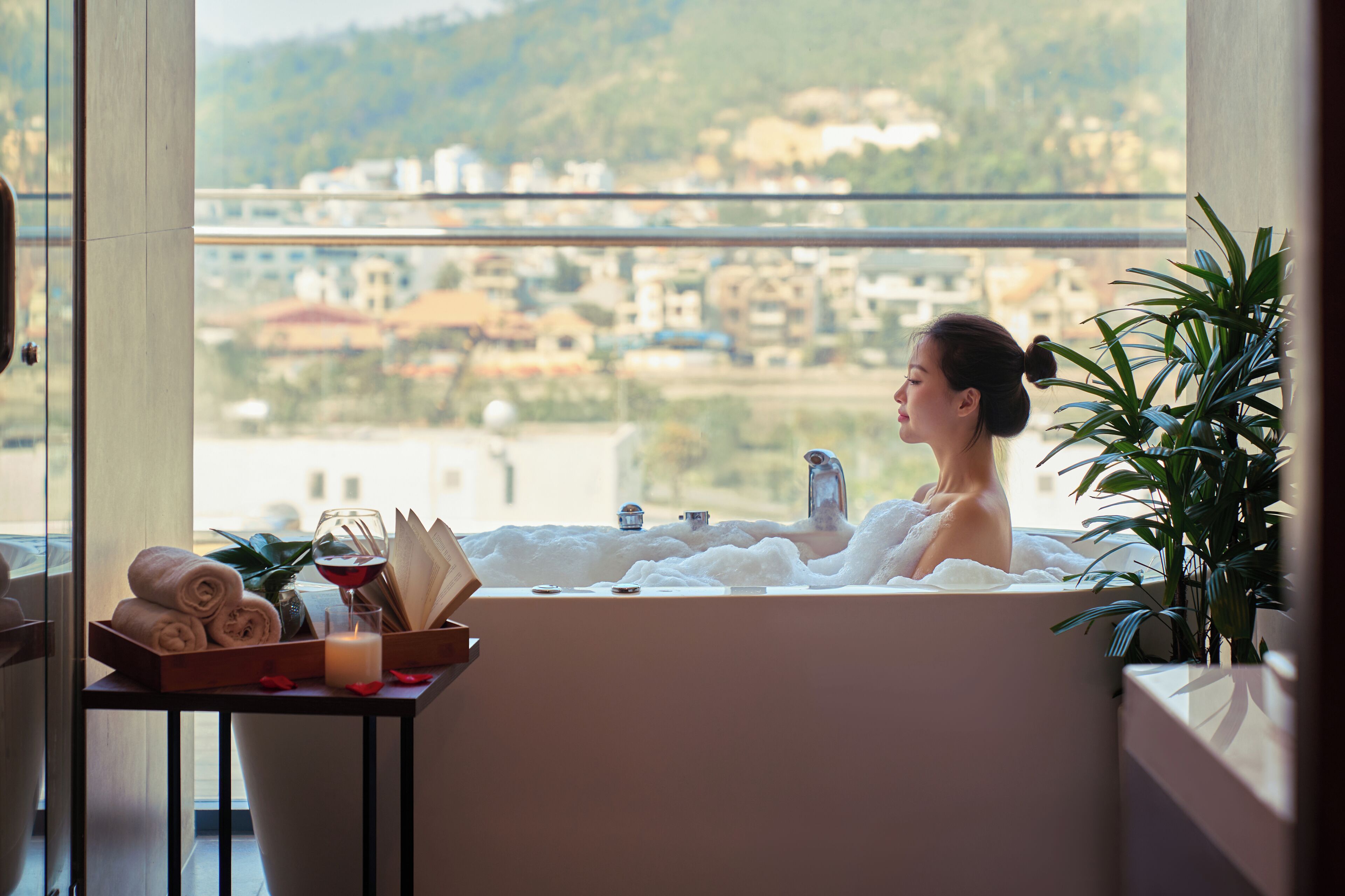 A woman in a bathtub with a city view, a towel rack, and a tray with towels and a glass of wine.