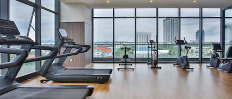 Une salle de sport spacieuse équipée de tapis de course, de vélos d’appartement et d’un rameur, avec de grandes fenêtres offrant une vue sur la ville.
