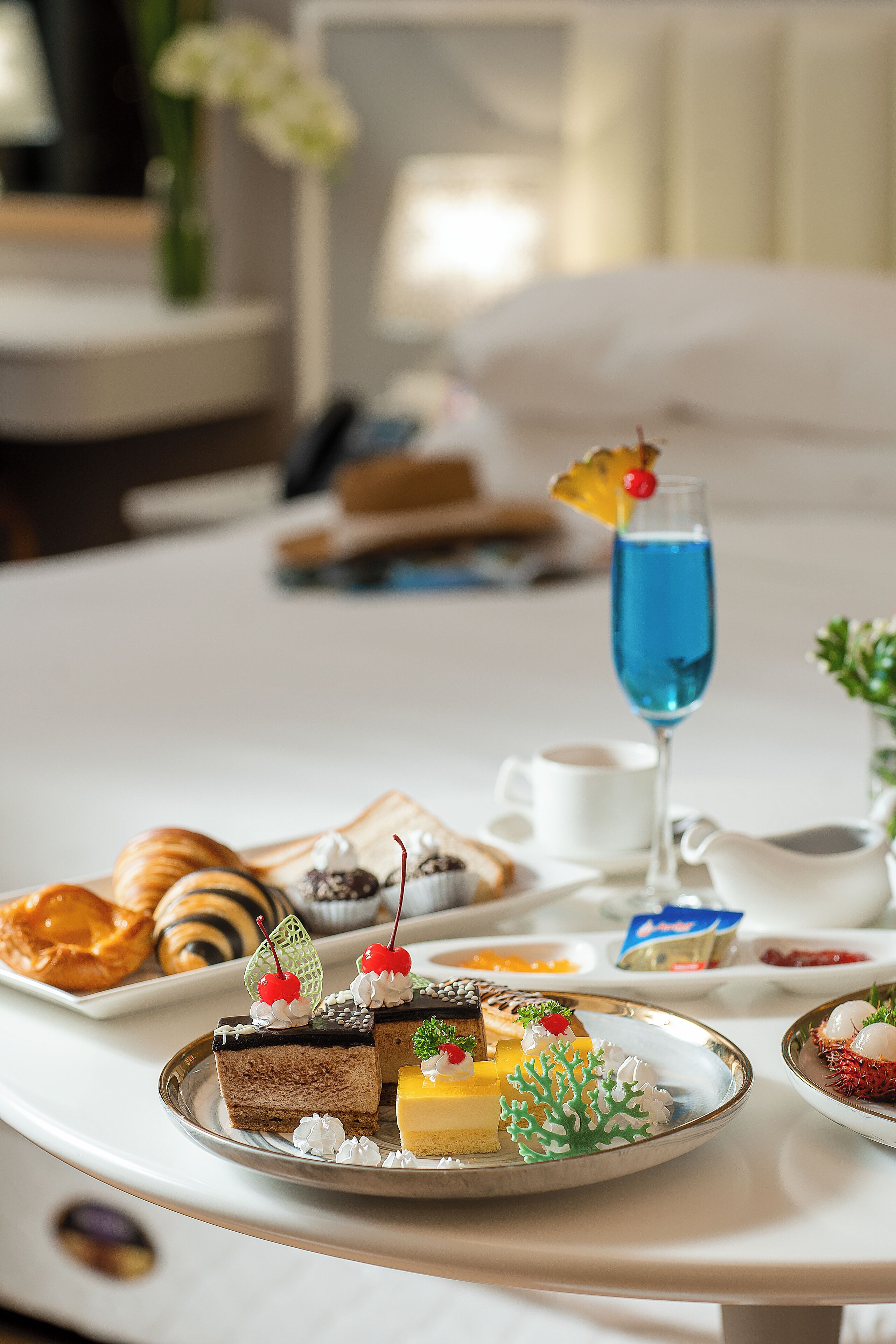 A breakfast tray with various pastries, a glass of blue beverage, and a small white pitcher on a hotel table.