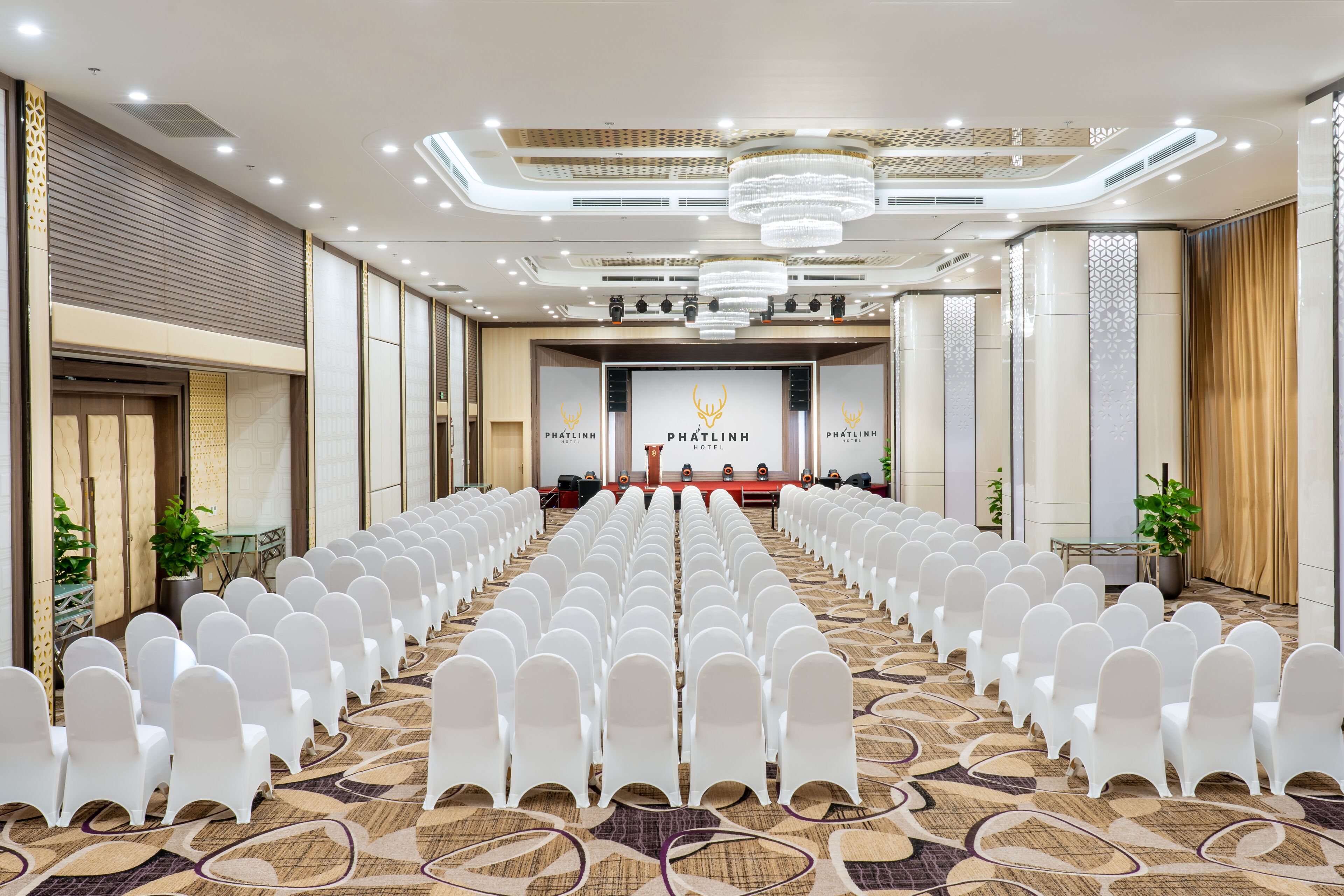 Une salle de conférence avec des rangées de chaises blanches, une scène avec un pupitre et un lustre.