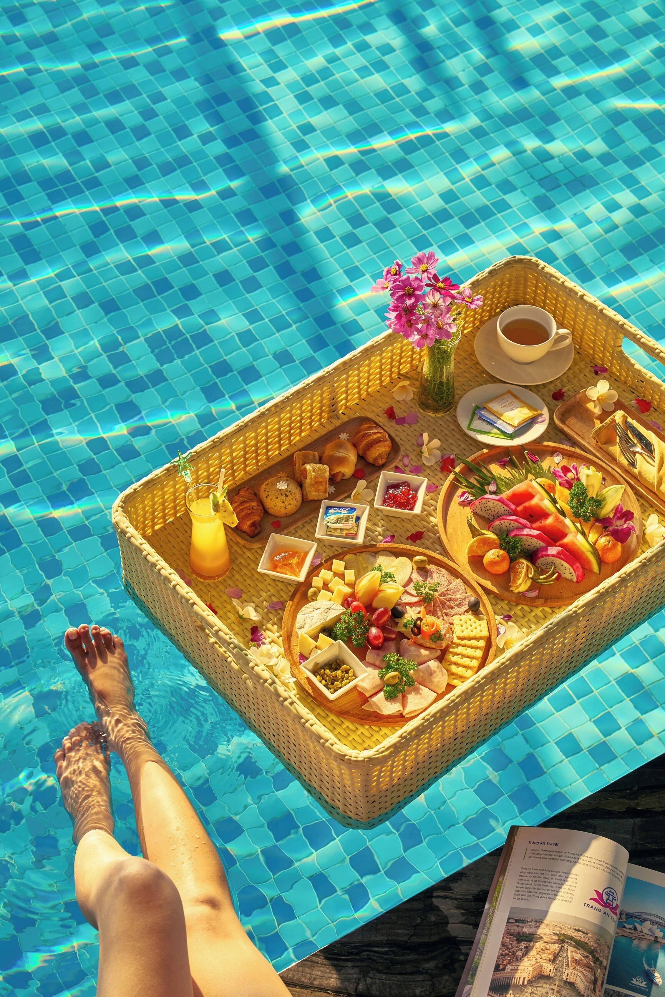 Desayuno junto a la piscina con una bandeja de mimbre que contiene comida, bebidas y flores.