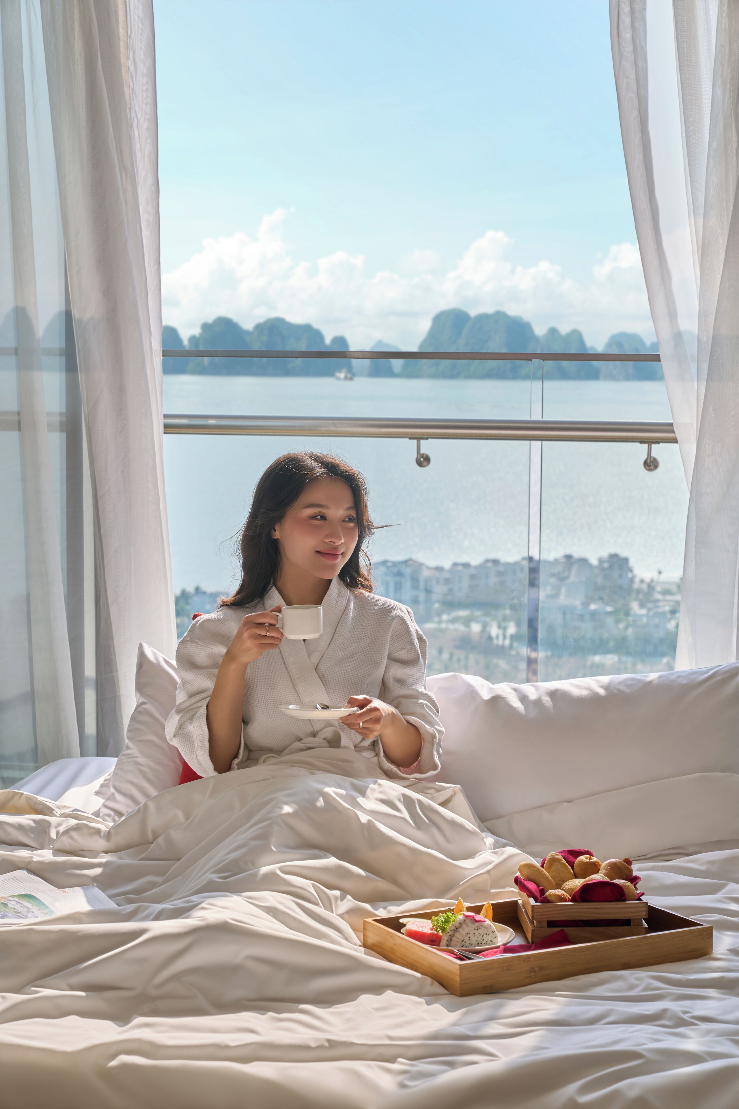 Une personne est assise sur un lit, avec un plateau de nourriture et une tasse, près d'une fenêtre donnant sur la ville et les montagnes.