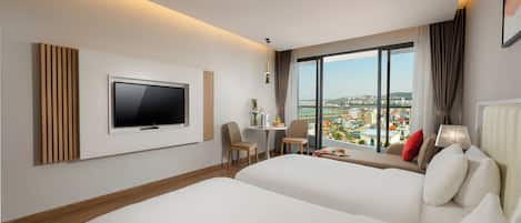 A modern hotel room with two beds, a TV, a desk with chairs, and a large window offering a city view.