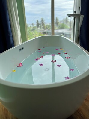 A bathtub with water and flower petals, a window with a view of greenery, and a curtain.