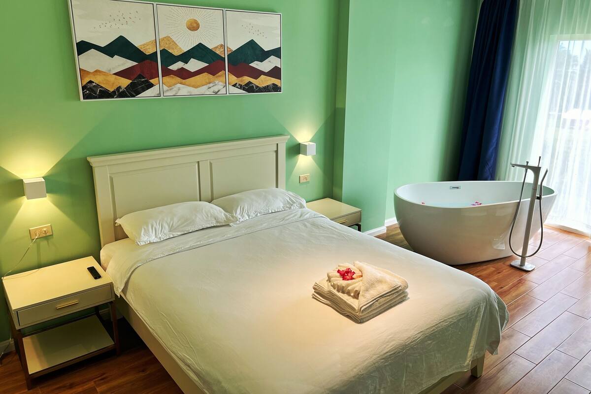 Ein ordentlich bezogenes Bett mit Handtuch und Rose, ein weißer Nachttisch mit Fernbedienung, ein Wandbild und eine große Badewanne.