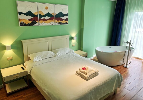 A neatly made bed with a towel and rose, a white nightstand with a remote, a wall-mounted artwork, and a large bathtub.