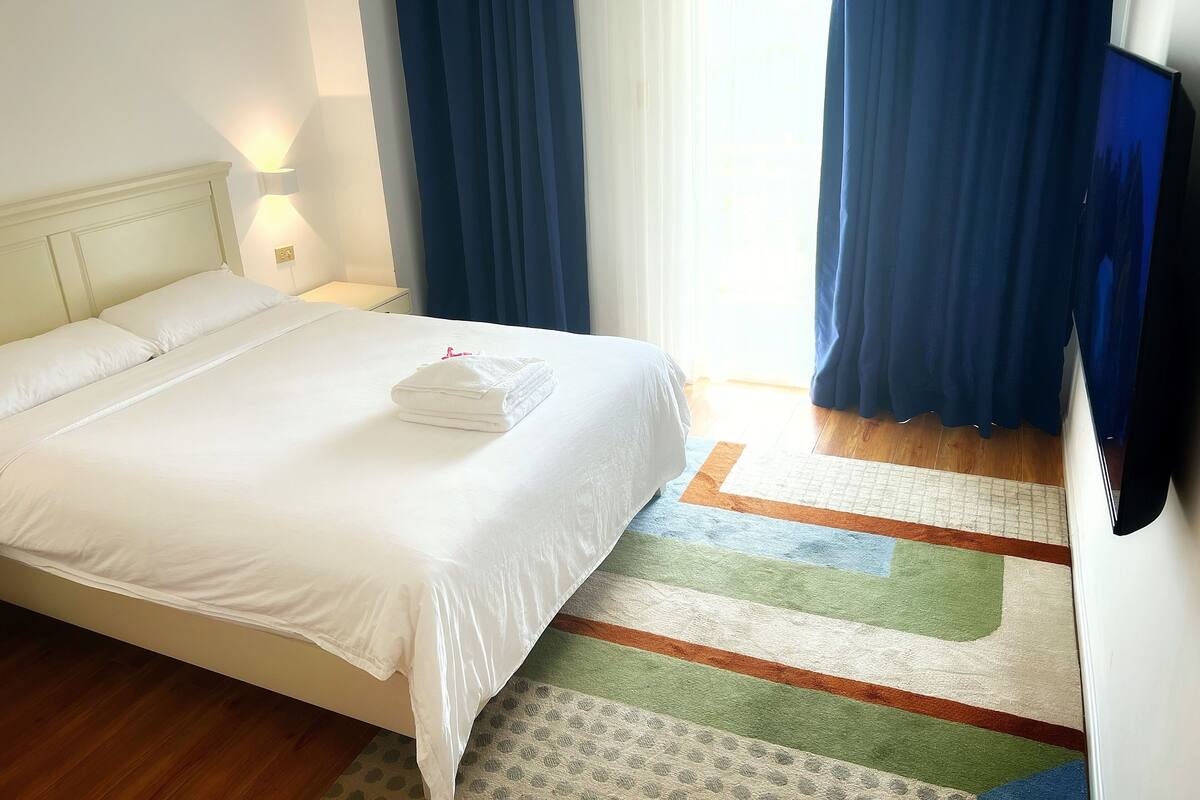 Ein Hotelzimmer mit einem ordentlich bezogenen Bett, einem Nachttisch mit Lampe, einem Fernseher und einem Fenster mit blauen Vorhängen.