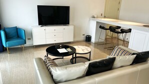 Una sala moderna con sofá, una silla azul, una mesa de centro, televisor y un área de cocina con taburetes.
