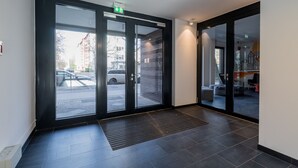 Un moderno vestíbulo de hotel con entrada por puerta de vidrio, zona de estacionamiento visible en el exterior y un área de descanso con sillas y mesas.