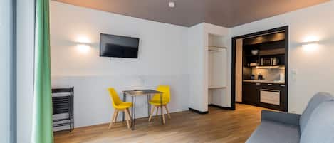 Een moderne woonkamer met een grijze bank, een eethoek met een kleine tafel en gele stoelen, een flatscreen televisie aan de muur en een keuken met een magnetron en kastjes.