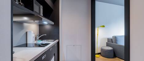 A modern kitchen with a built-in oven, a sink, and a countertop. There is a small window above the sink and a door leading to a living area with a sofa and a lamp.