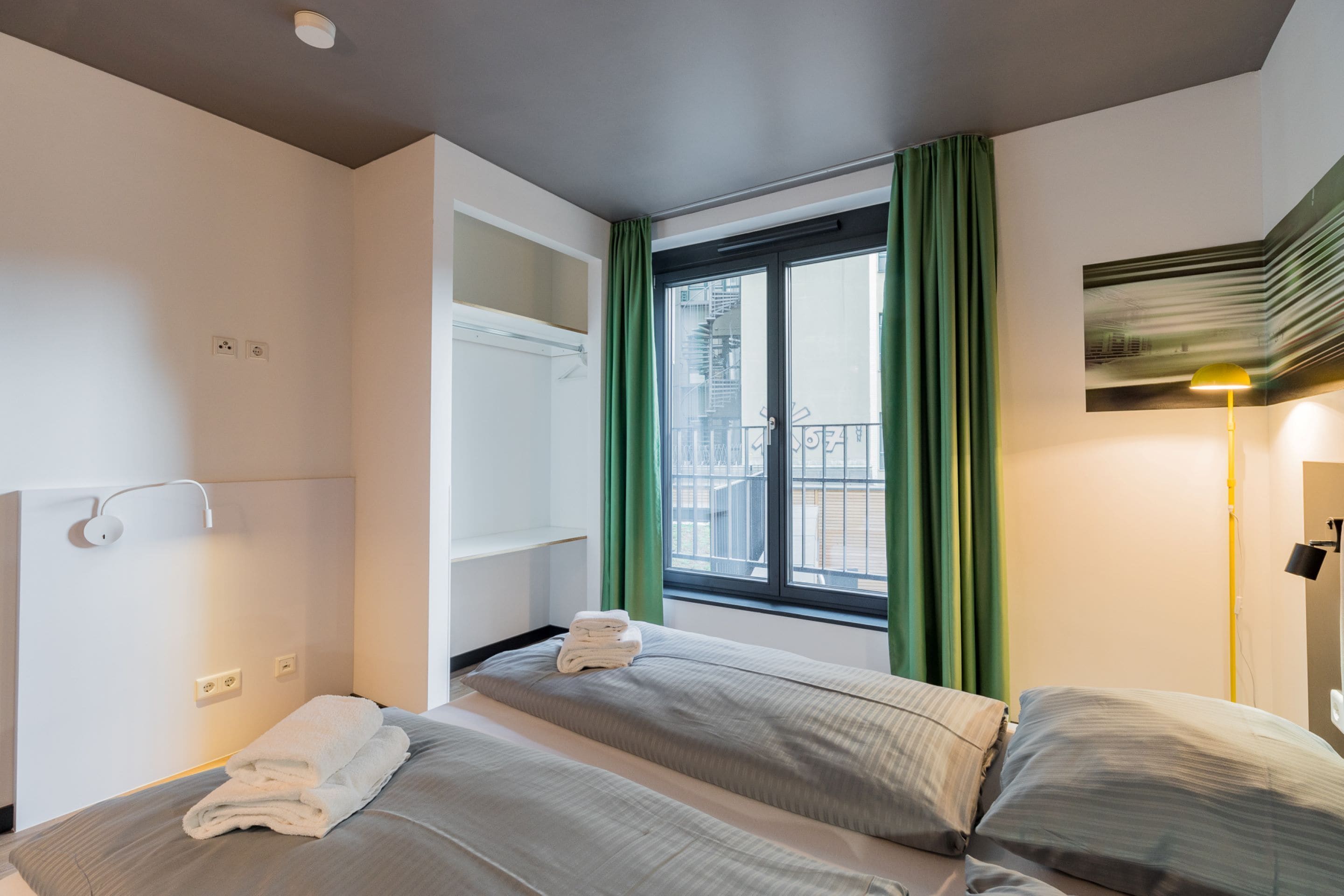 Une chambre d’hôtel avec deux lits, une fenêtre avec des rideaux verts, un balcon et une lampe fixée au mur.