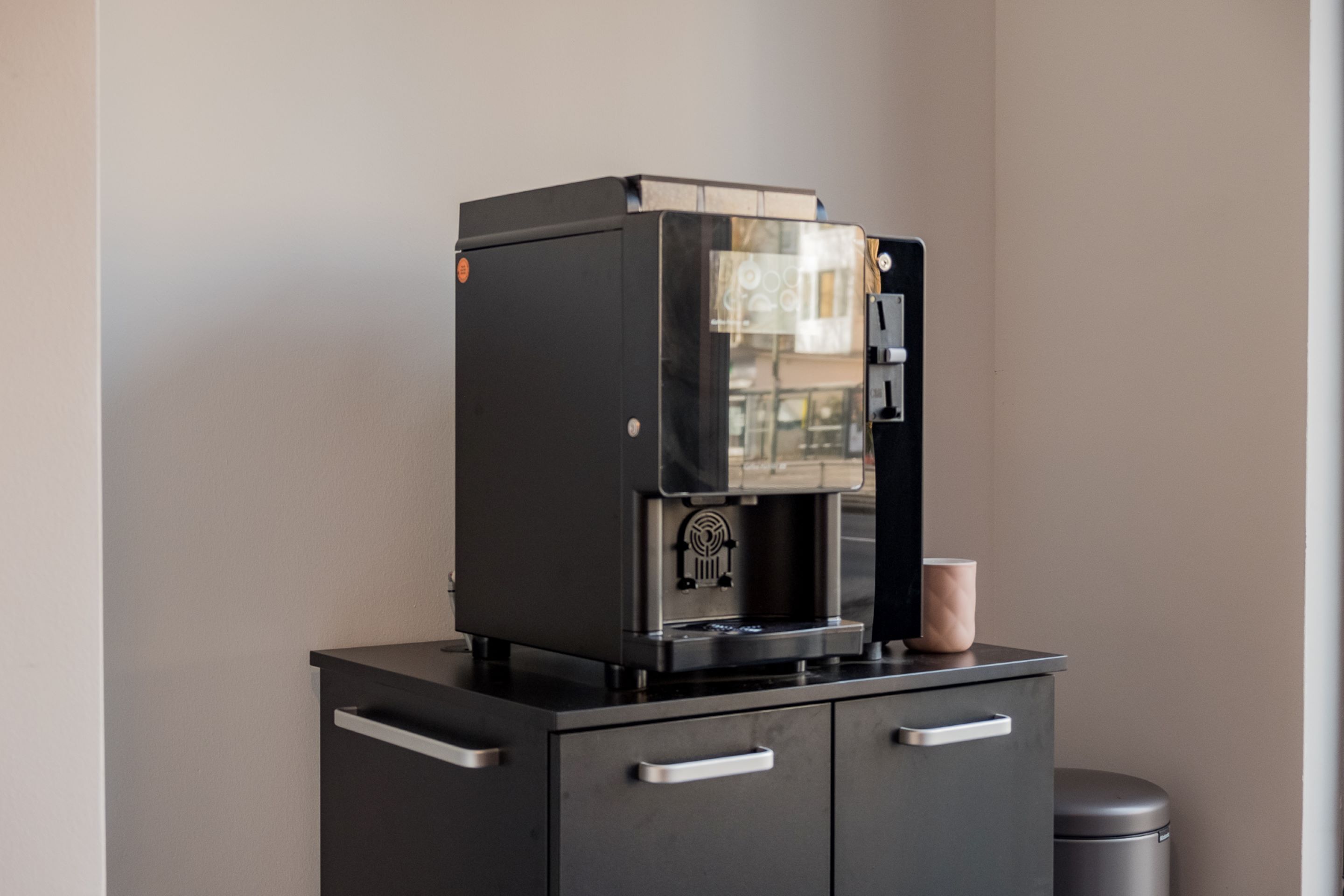 A coffee machine on a black cabinet with drawers, a cup holder, and a trash can in the background.