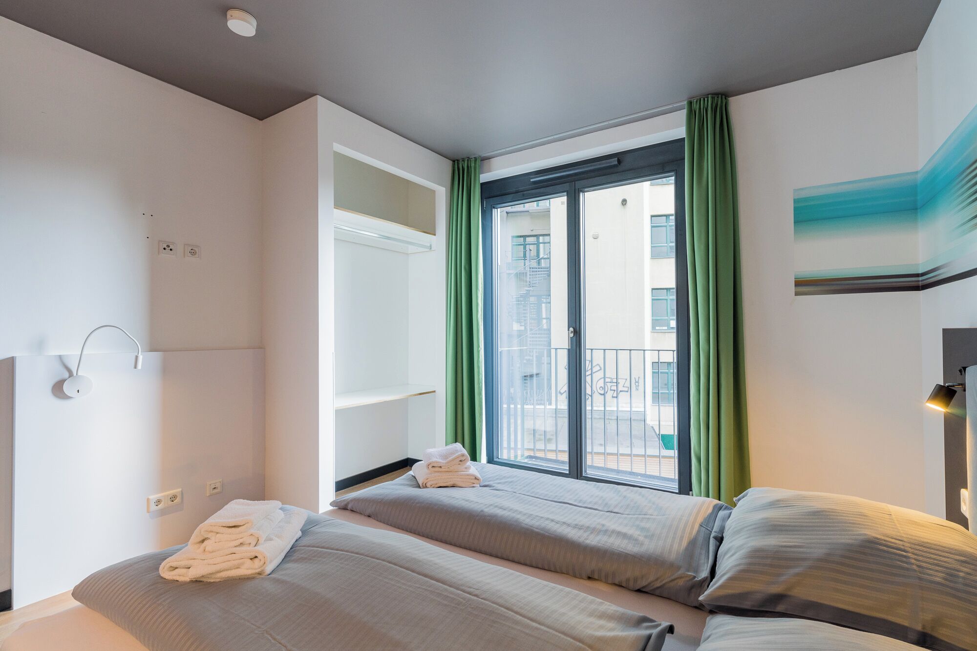 A hotel room with two beds, a balcony with green curtains, a view of buildings, and a painting on the wall.