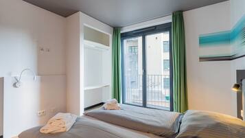 A hotel room with two beds, a balcony with green curtains, a view of buildings, and a painting on the wall.
