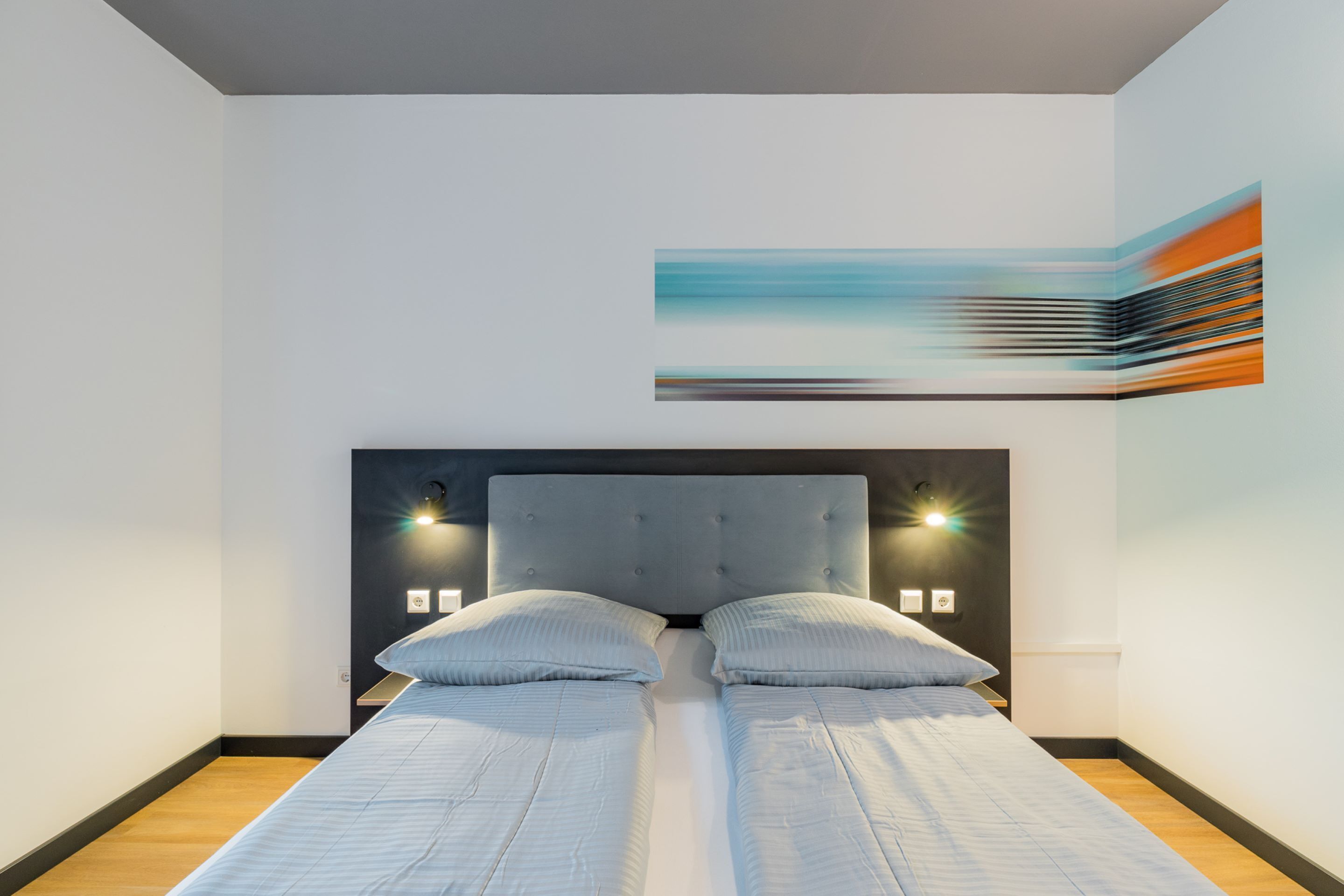 A modern bedroom with a large bed, two wall-mounted lights, and a contemporary abstract painting.