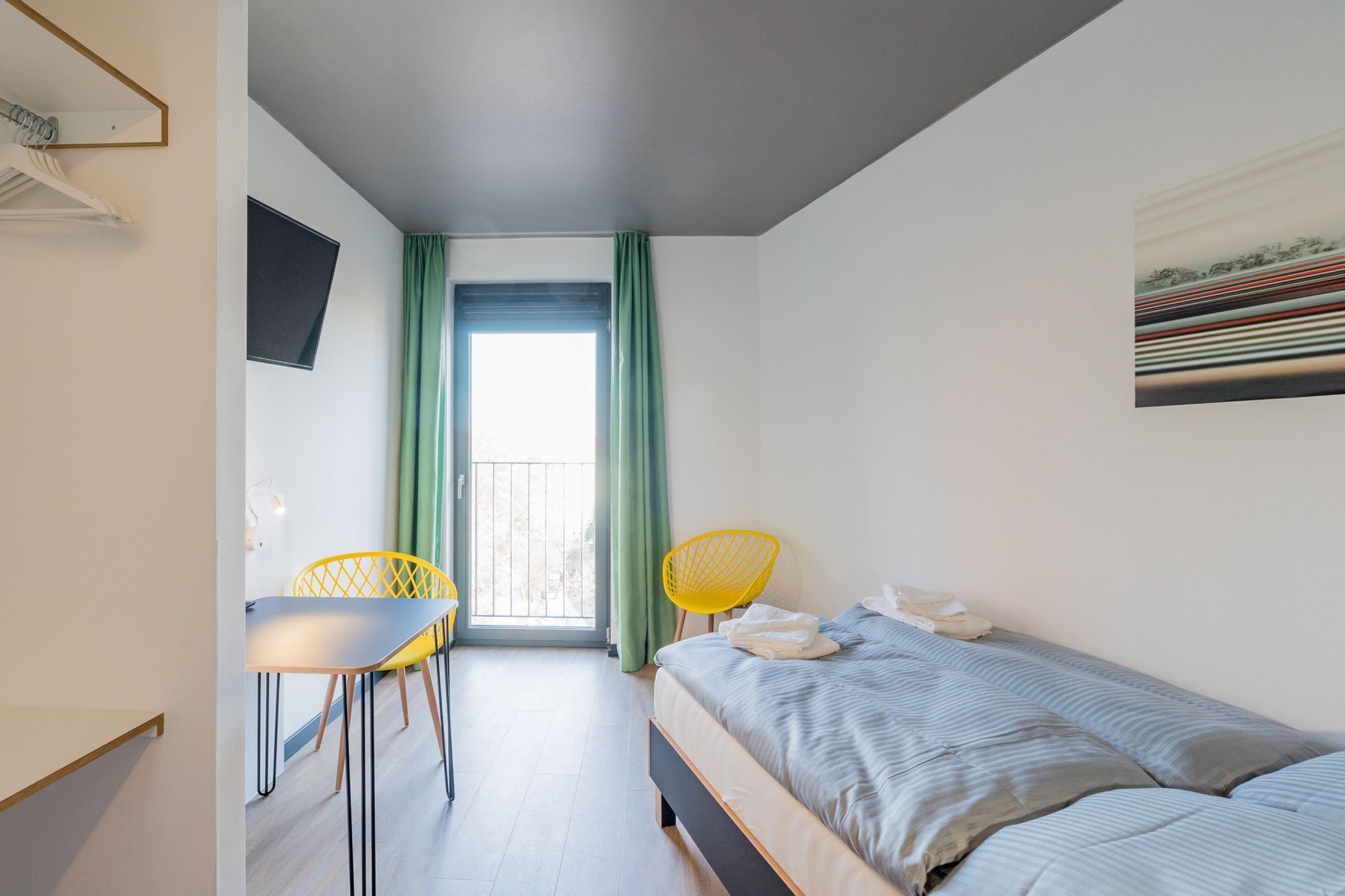 A bedroom with a bed, a desk, two chairs, a TV, and a balcony with curtains.