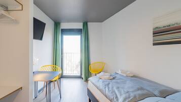 A bedroom with a bed, a desk, two chairs, a TV, and a balcony with curtains.