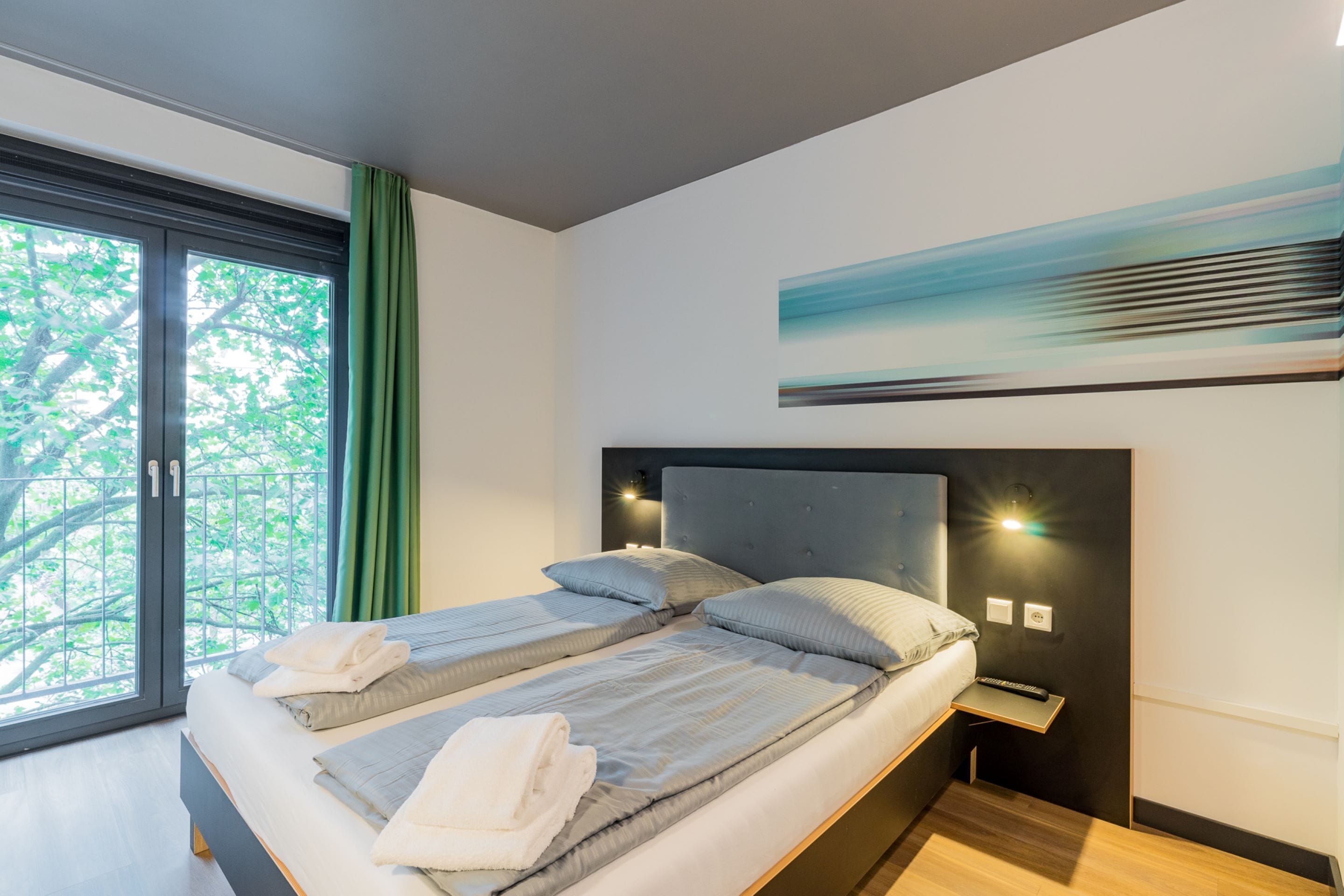 A modern bedroom with two beds, a large window with a view of trees, and a painting on the wall.