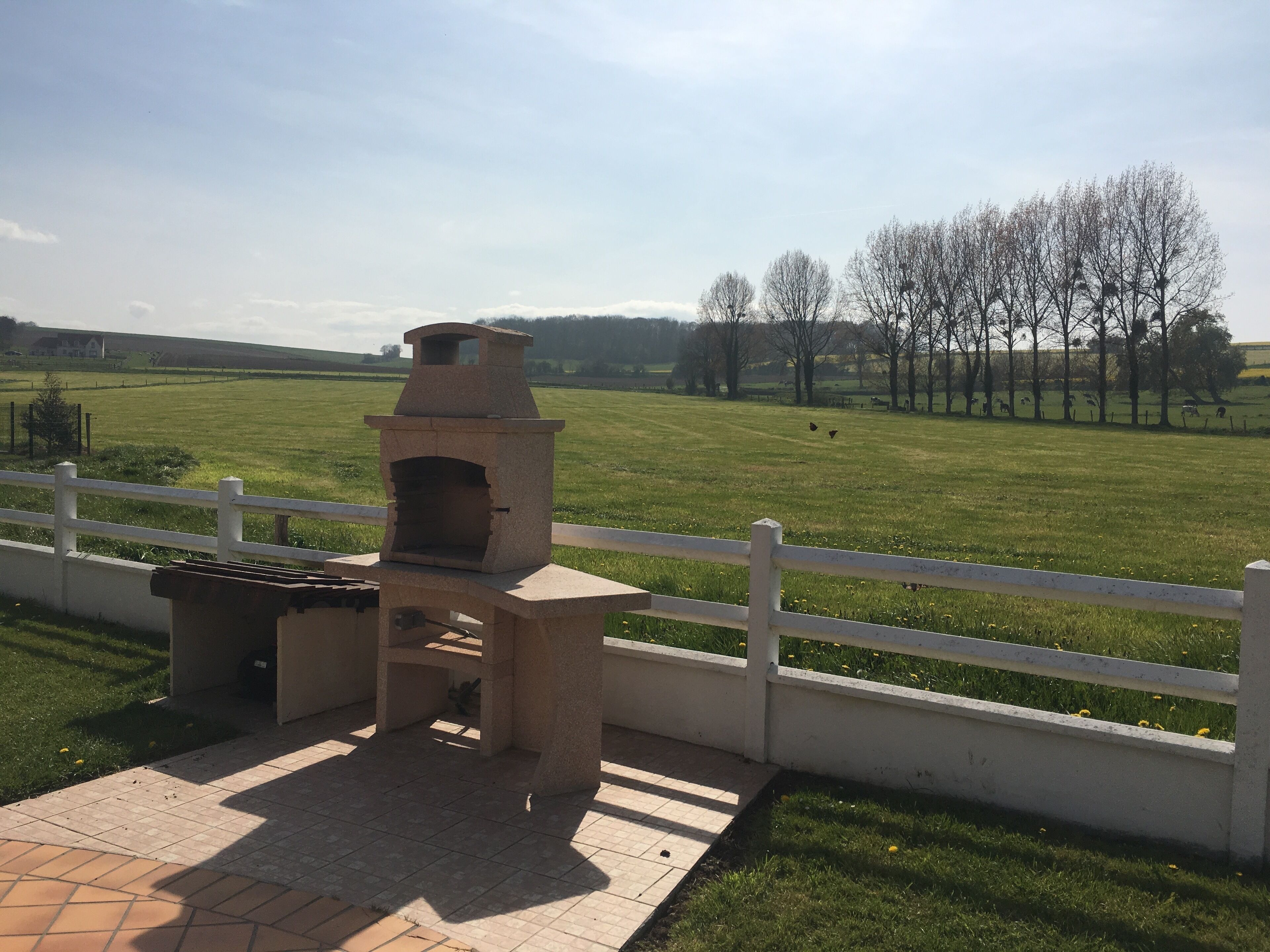 A stone outdoor kitchen with a chimney, surrounded by a white fence and a paved patio area.