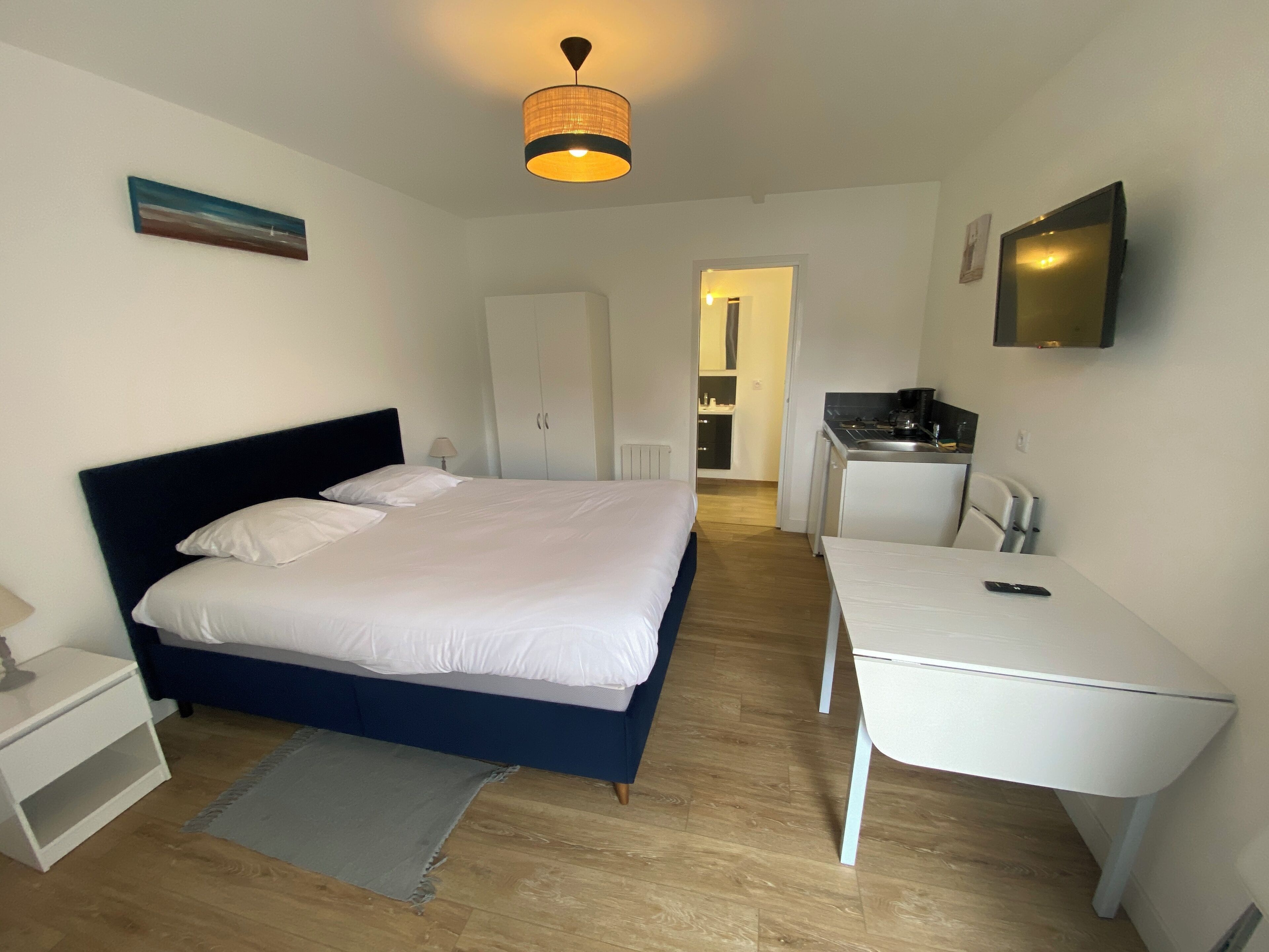 Une chambre à coucher avec un lit, une table de chevet et un bureau avec une chaise. On y trouve un luminaire au plafond, un tableau accroché au mur et une porte donnant sur une salle de bain.