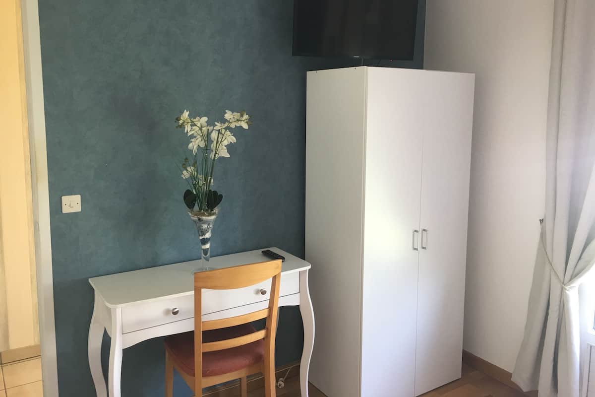 Een kamer met een wit bureau, een houten stoel, een witte kledingkast en een flatscreen televisie die aan de muur is bevestigd.