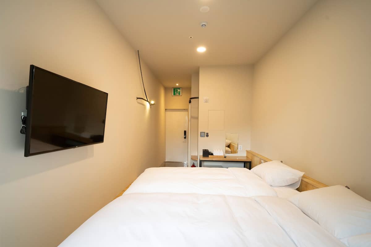 A hotel room with a large bed, a TV mounted on the wall, a bedside table with a mirror, and a door leading to a bathroom.