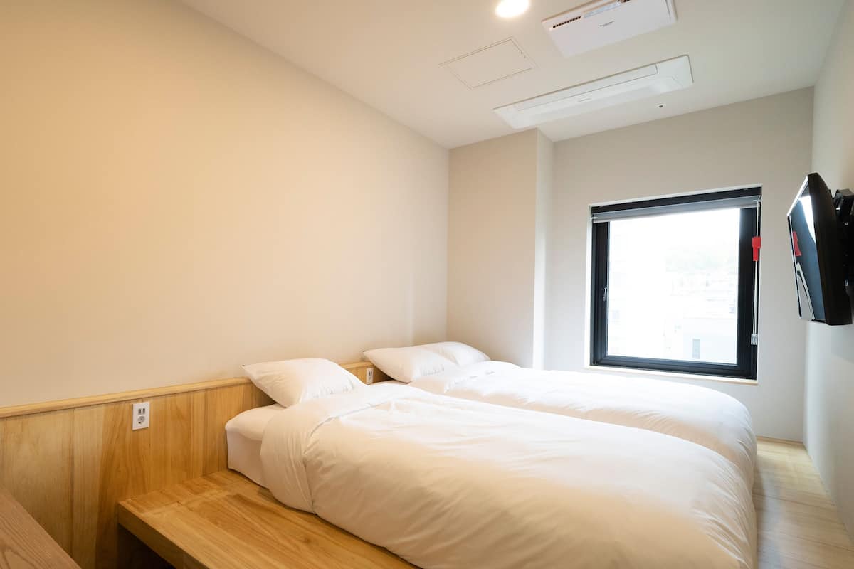 A hotel room with two single beds, a wooden bench, a window with blinds, and a mounted TV.