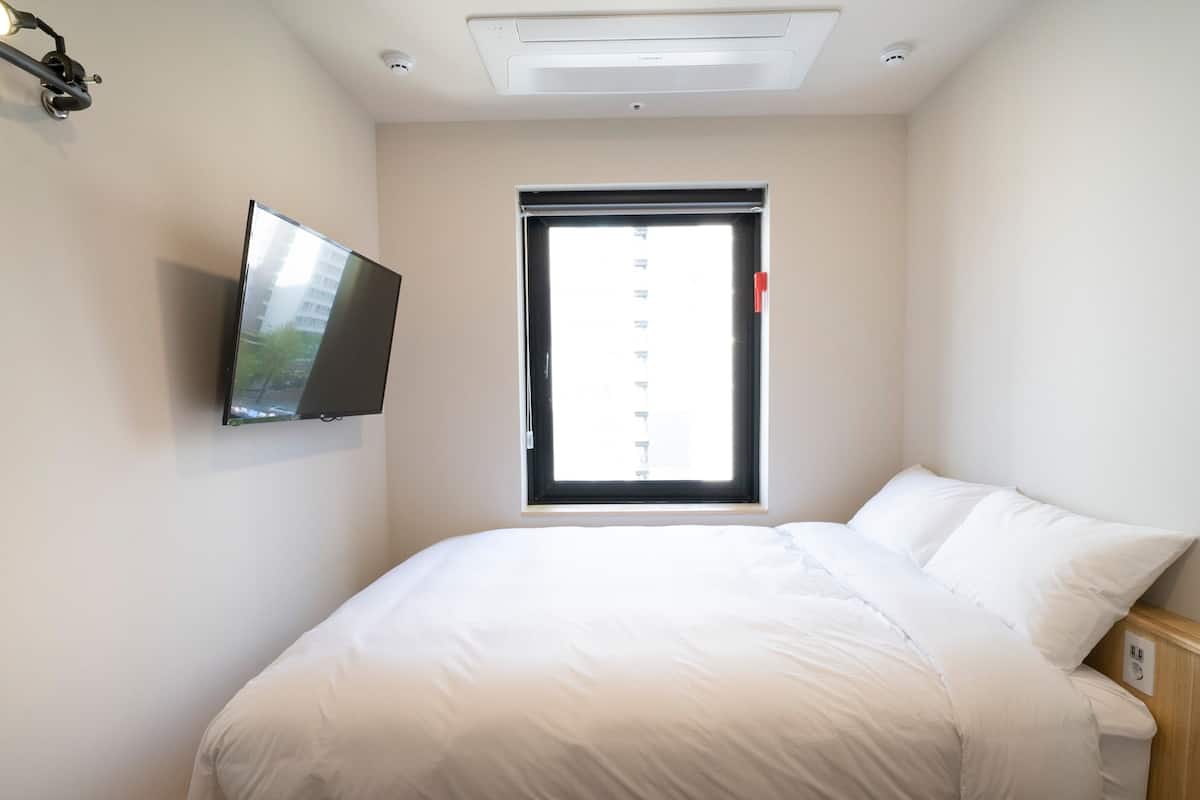 A bedroom with a bed, a television mounted on the wall, a window with blinds, and a ceiling light.