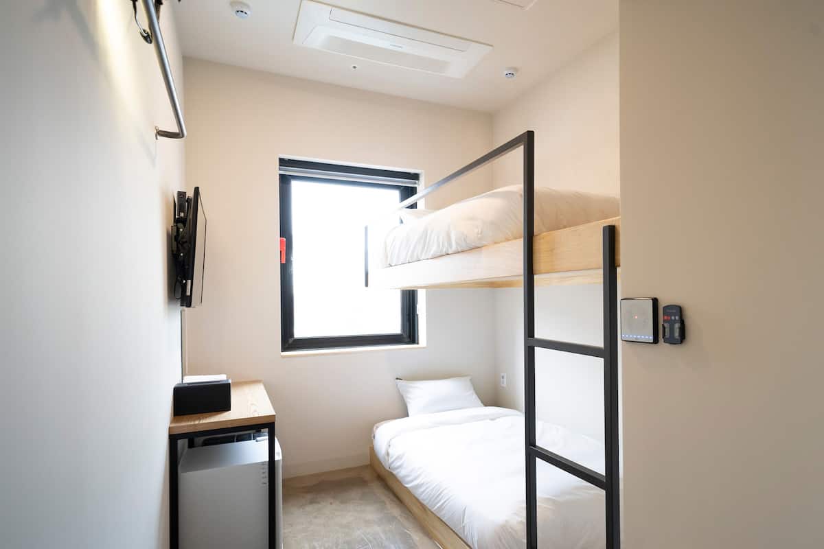 A compact dormitory room with bunk beds, a desk, a small kitchenette, and a window providing natural light.