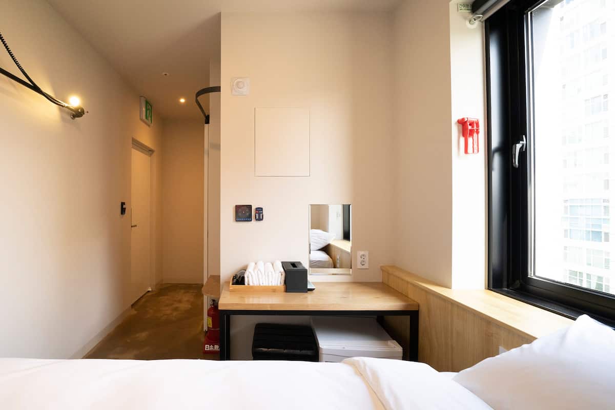 A hotel room with a bed, a desk, a chair, a fire extinguisher, a window, and a door.