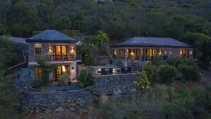 A two-story stone house with a balcony and outdoor seating area, surrounded by lush vegetation.