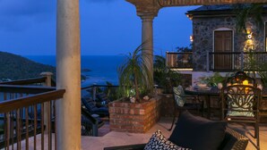 A stone building with a balcony overlooking the ocean, featuring outdoor furniture and a brick planter.