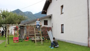 Um parquinho com escorrega e equipamentos de escalada, uma pessoa em uma escada e uma casa ao fundo.