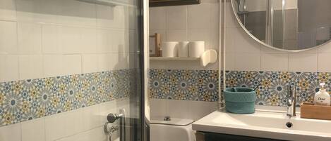 Un baño con cabina de ducha de vidrio, inodoro, lavabo con espejo y repisa con artículos de aseo.