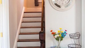 A staircase with a dark wooden handrail, a white wall with a round clock, and a small white table with a vase of flowers and magazines. - Charming Suite in the Heart of Charleston (Charleston)