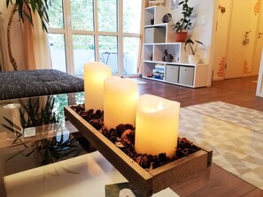 A modern living room with a glass coffee table, three lit candles in a wooden box, a sofa, and a bookshelf with decorative items.