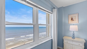 A room with a large window overlooking a beach, a white dresser with a lamp, and a framed picture on the wall. - Stunning Oceanfront Condo with Spectacular Views of NSB (New Smyrna Beach)