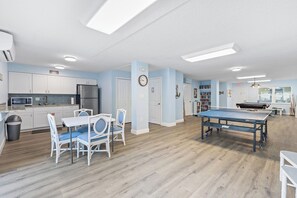Un vaste espace de vie comprenant une table de ping-pong, une cuisine avec des meubles blancs et un coin repas avec une table blanche et des chaises bleues.